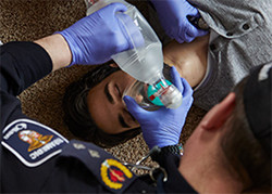 Emergency Medical Services person kneeling next to person lying on the ground and holding oxygen mask over the person's face