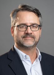 Professional headshot photo of Ian Ketcheson, wearing a light checkered shirt and back blazer against a neutral gray background.