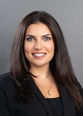 Professional headshot photo of Joelle Raffoul, smiling and wearing a black blazer against a neutral gray background.