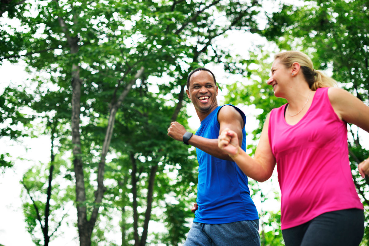 Male and female in exercise gear smiling at each other while running in a green neighbourhood
