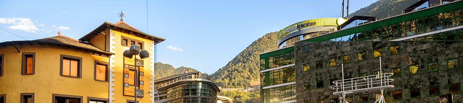 A yellow building and glass building sit in front of green, tree covered hills in Andorra.