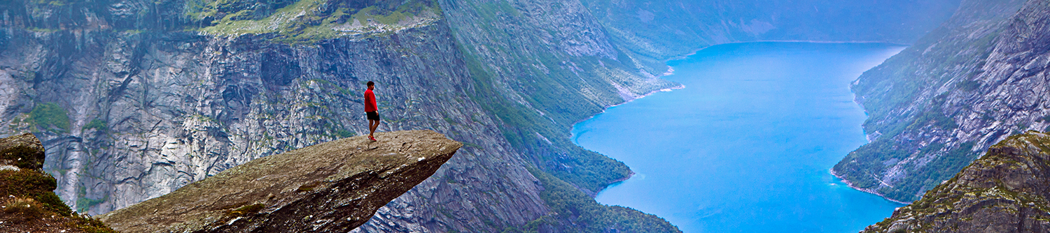 A person stands on an outlook in the mountains over a body of turquoise blue water in Norway