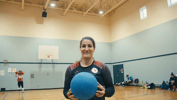 In a gym, Maryam holds a goalball and smiles.