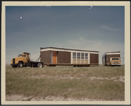 Photographie en couleur présentant deux bâtiments préfabriqués transportés par deux camions.