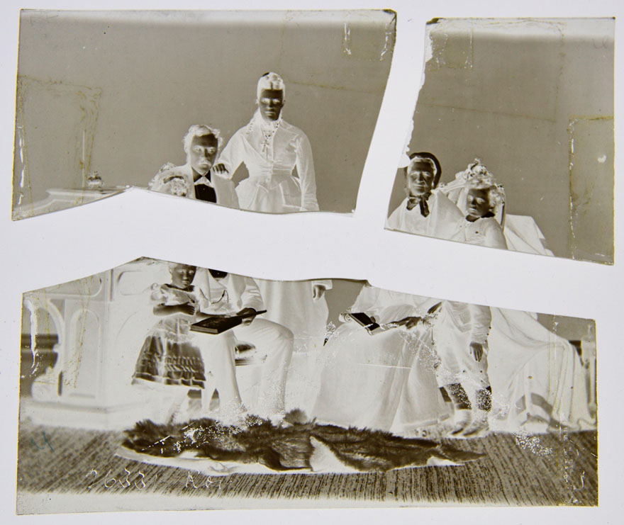 A glass plate negative broken into three pieces. It is shown as a negative. The image is of a family portrait: a small child is standing while reading a book with her father as he is seated, standing beside him, his wife, seated beside her an elderly woman and beside her a small boy is standing.