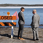 Prince of Wales and another person speaking next to a closed road sign, in front of water
