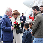Prince of Wales and Duchess of Cornwall smiling and talking children and adults on tarmac with a plane and officials in the background.