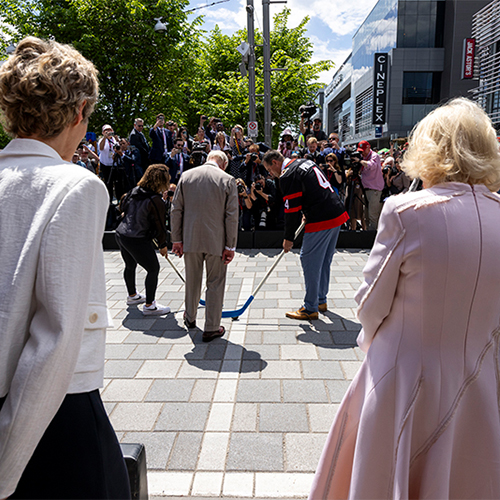 Le Roi Charles III se tient debout, tenant une rondelle de hockey, entre deux personnes munies de bâtons de hockey.