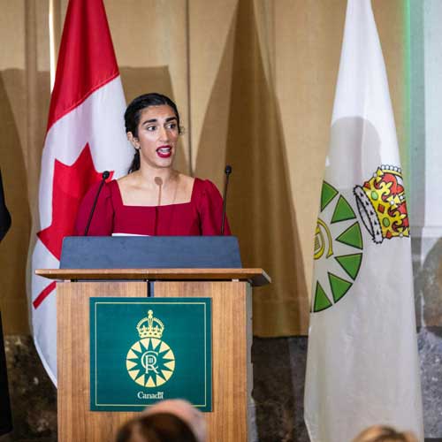 A woman speaks at a podium to a seated audience. The National Flag of Canada and a flag with the Canadian Coronation Emblem are behind her.