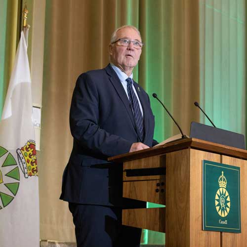 A man speaks from a podium. To his right is a flag with the Canadian Coronation Emblem.