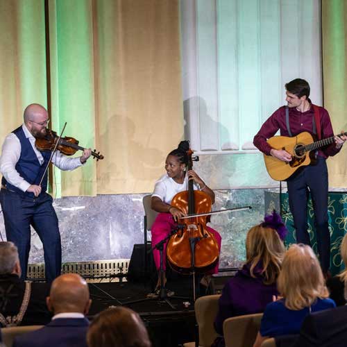 A band consisting of a fiddler, cellist and guitarist performs in front of an audience.