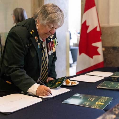 A woman in a military uniform with decorations signs a book placed on a table.