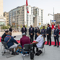 A group of dignitaries stand on a red carpet watching a drumming performance. There are Canadian flags and city buildings behind them. The four drummers are seated.