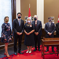Un groupe de dignitaires, ainsi que la gouverneure générale, sont debout derrière une petite table en bois. Tous sont masqués.