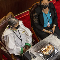An Inuk Elder dressed in traditional clothing sits next to a woman in a black jacket and blue blouse. In front of them is a small table with a lit qulliq, a traditional Inuit oil lamp. They are both masked.