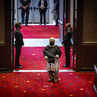 An Inuit drummer in traditional clothing, carrying a drum and stick, walks through the doorway to the Senate chamber. The carpet is red with stylized maple leaves in yellow and blue.