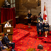 An Algonquin Elder is speaking at the podium. She wears a white top. To her right seated is the Governor General Designate and her spouse. The Senate carpet is red. We see the Canadian flag and the Senate throne in the background.