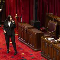 A performer is standing, singing into a handheld microphone. She is wearing a black suit and white shirt. We see the red carpet and wood seats of the Senate Chamber. Two people are seated to her right and listening to her performance.