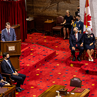 Un homme portant un complet bleu/gris parle derrière un podium. À sa droite, la gouverneure générale désignée, son conjoint et une autre personne sont assis. À l'avant se trouvent le premier ministre et sa conjointe. À l'arrière-plan, on voit un drapeau canadien et le trône du Sénat.