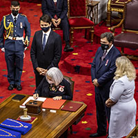 The Governor General is seated and is signing a book. The Prime Minister and a uniformed military member stand to her left. A woman and a man stand on her right. All are masked. We see the collars of office on the table on blue pillows.