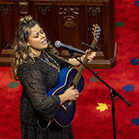 Une interprète, vêtue d'une robe noire, joue d'une guitare bleue et chante dans un microphone sur pied. Nous voyons le tapis rouge du Sénat et des sièges.