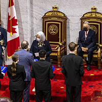We see the Governor General in the centre of the photo with several people standing in front of her. She is masked. Behind her we see her spouse sitting on one of the two thrones, a uniformed officer and a Canadian flag.