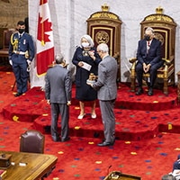 A wide-angle shot of the Governor General with two men in grey suits standing in front of her. She is wearing a navy dress and is masked. Behind her, we see her spouse sitting on one of the two thrones, and two Canadian flags on either side.