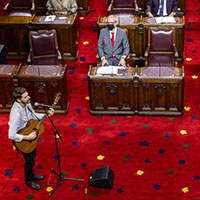 Plan large d'un interprète qui porte une chemise de couleur claire. Il joue de la guitare et chante dans un microphone sur pied. On voit des gens qui regardent depuis les sièges du Sénat.