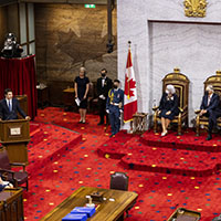 A wide-angle shot of the Governor General and her spouse sitting on the thrones in the Senate. They are both masked. The Senate carpet is red and the wall behind them is white. There are two Canadian flags on either side. We see the Prime Minister of Canada speaking at the podium on their left.