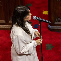 Une interprète portant un chemisier blanc chante dans un microphone sur pied. Derrière elle, nous voyons le tapis rouge du Sénat et des sièges.