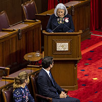 La gouverneure générale parle au podium. Elle porte une robe marine avec des broderies à l'encolure. Elle porte ses honneurs. On voit le premier ministre et sa femme en avant plan.