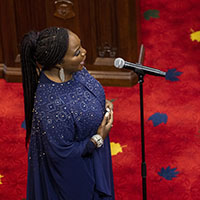 Une interprète vêtue d'une longue robe bleue marine chante dans un microphone sur pied. Derrière elle, nous voyons le tapis rouge du Sénat et des sièges.