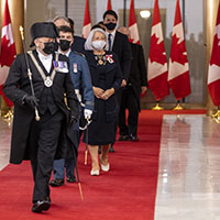 The Usher of the Black Rod, dressed in his traditional black costume and carrying a black and golden rod, leads the Governor General and the group of dignitaries on the red carpet. Everyone is masked. We see Canadian flags behind them.