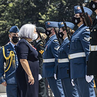 La gouverneure générale est debout à l'extérieur et passe en revue des membres des Forces armées canadiennes en uniforme. Tout le monde est masqué.
