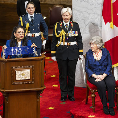 An Algonquin Elder speaks at a podium. The governor general designate, the Chief of the Defence Staff, and an aide-de-camp are nearby.