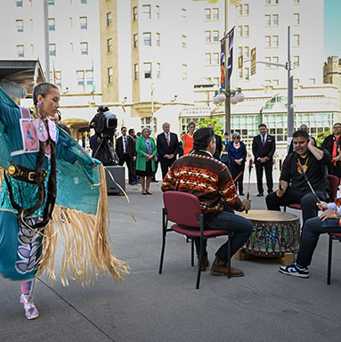 An Algonquin dancer in colourful regalia dances next to a group of four seated men beating a drum.