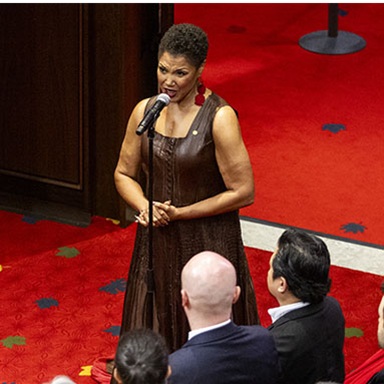 A woman stands and sings into a microphone. There are rows of guests standing on either side.