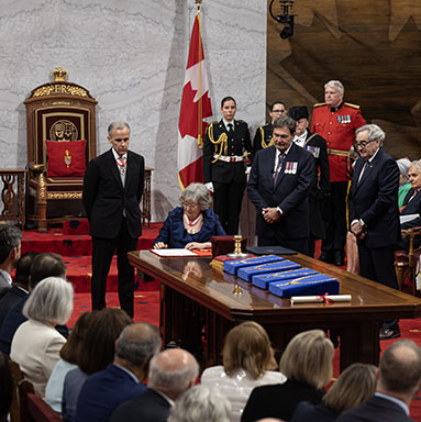 The Governor General Designate sits at a table in the Senate Chamber and signs a document. Government dignitaries are standing nearby.