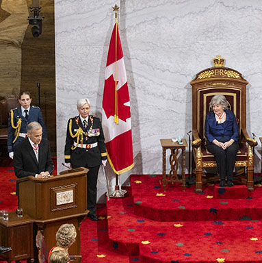 The Prime Minister speaks behind a podium. The Governor General is seated on the Throne. Three military personnel are standing nearby.