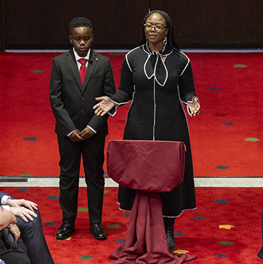 A boy stands next to a woman who is speaking behind a draped podium. There are rows of seated guests on either side.