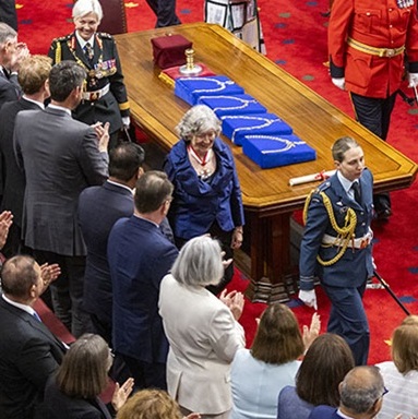 The Governor General escorted by military personnel walks by a table with ceremonial collars between rows of standing guests.