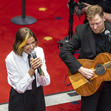 A performer is standing and singing into a handheld microphone. A man is playing the guitar next to her.