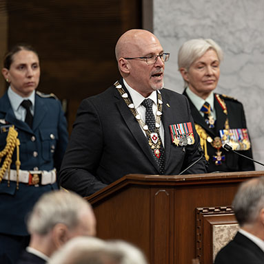 The Secretary to the Governor General speaks at a podium. The Chief of the Defence Staff and an aide-de-camp are standing in the background.