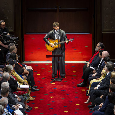 A man stands singing into a microphone while playing the guitar. There are rows of seated guests on either side.