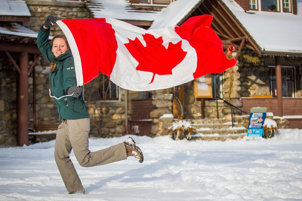 A young woman smiling while running in the snow holding a large Canadian Flag.