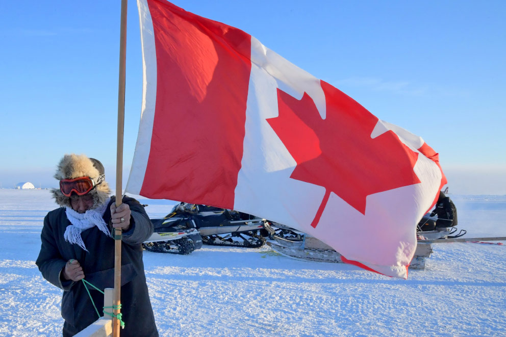 A man dressed very warmly tying the pole of a large Canadian Flag to a post. Snow, a blue sky and a snow mobile can be seen in the background.