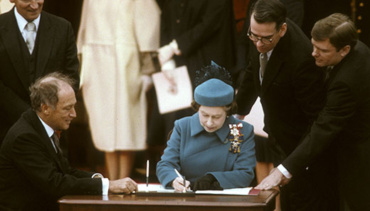 Photographie en couleur de la Reine signant un document officiel. L’ancien premier ministre Pierre Elliott Trudeau est assis à sa droite