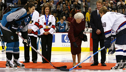 The Queen standing on a red carpet, about to drop a hockey puck. Wayne Gretzky is behind as two hockey players are ready to catch the puck