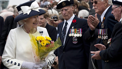 The Queen is carrying a bouquet of flowers. She is greeting a group of veterans dressed in military attire