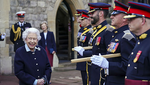 The Queen is smiling, facing a row of officers in military uniforms. Behind her is a stone building with an arched doorway